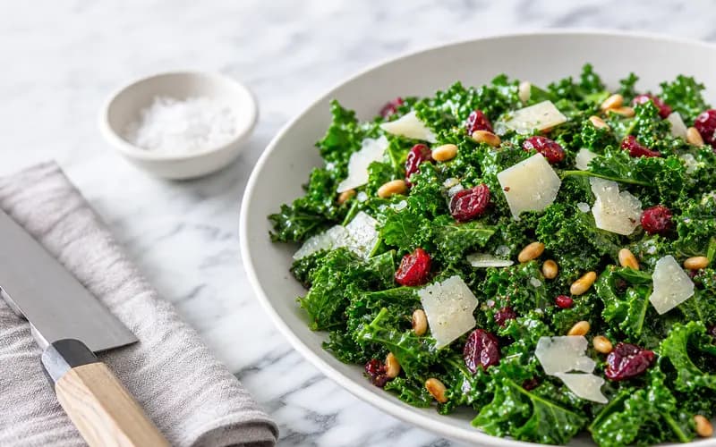 Salade de kale vert foncé massé, cranberries rouges, copeaux de parmesan et noix dans un grand bol blanc