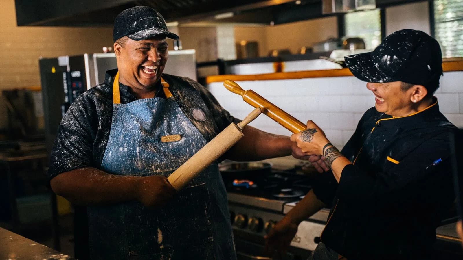 Deux chefs complices en cuisine, riant ensemble autour de rouleaux à pâtisserie et de farine, symbole du partenariat éditorial MonCoachGourmand